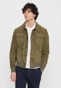 Giacca in suede verde oliva con due tasche sul petto, chiusura a bottoni e orlo a costine. Indossata sopra una camicia bianca, esposta su uno sfondo semplice.