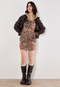 Chaqueta de bomber de cuero marrón sobre un vestido estampado en beige y rojo con diseños de mandala. Botas negras hasta la rodilla con calcetas claras.