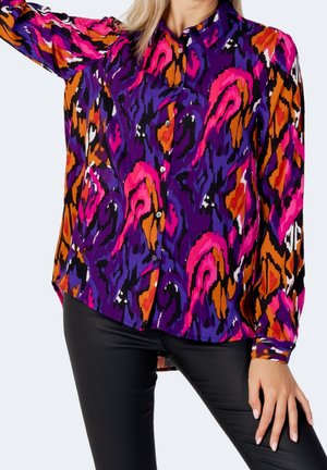 Kvinde iført en livlig skjorte med knapper med abstrakte lilla, pink, orange, sorte og hvide mønstre kombineret med sorte bukser.