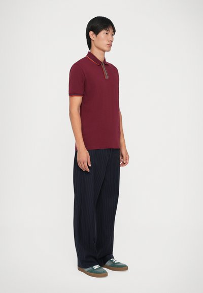 Μπορντό πόλο μπλούζα με αντίθετο ριγέ γιακά, συνδυασμένη με σκούρο παντελόνι με ρίγες και πράσινα αθλητικά παπού�τσια με καουτσούκ σόλα.