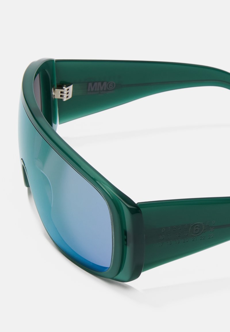 Occhiali da sole verdi con un design elegante a fascia e lenti blu sfumate; presentano un brand MM6 prominente e indicatori di taglia visibili.