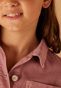 Roze corduroy shirt met een omgeslagen kraag, zilveren knoopsluiting en textuurstof. Korte bruine haren zichtbaar, samen met een klein oorbelletje.