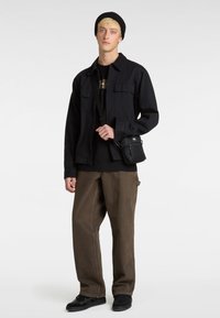 Sort denimjakke, mørk grafisk t-shirt, brune brede bukser, sort beanie og lille sort crossover-taske, båret med sorte sneakers.