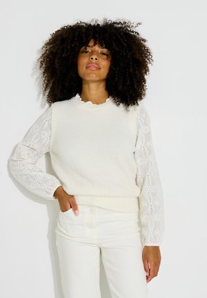 Femme aux cheveux bouclés portant un chemisier blanc brodé à manches longues sous un pull sans manches blanc, posant avec une main dans la poche.
