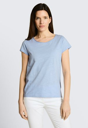 Kobieta o prostych ciemnych włosach, ubrana w jasnoniebieską koszulę z krótkim rękawem i białe spodnie, stojąca na jednolitym jasnym tle.