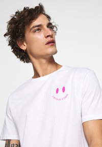 White cotton t-shirt with a round neckline featuring a pink smiley face and text "A BETTER ME IS COMING" in pink at the chest.