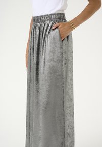 Jupe argentée métallique en tissu lisse et texturé. Dispose d'une taille élastique et de poches latérales. Design droit, longueur au sol.