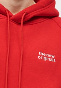 Sudadera con capucha roja con cordones delanteros y texto bordado en blanco "the new originals" en el área del pecho izquierdo, usada por una persona.