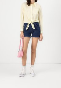 Chemise boutonnée jaune pâle nouée à la taille, associée à un short en denim foncé. Des baskets blanches et un petit sac à main rose complètent le look.
