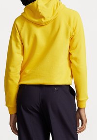 Ljusgul hoodie med en mjuk textur och justerbar huva, kombinerad med mörka byxor som har en knappdetalj vid bakfickan.