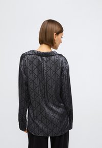 Cămașă cu mâneci lungi dintr-un material gri închis, cu un model geometric negru. Închidere cu nasturi la spate, croială lejeră și textură fină.