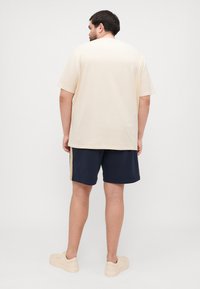 Beige t-skjorte med kort erm, løs passform, parret med marineblå shorts med sidestriper. Lyse sneakers fullfører antrekket.