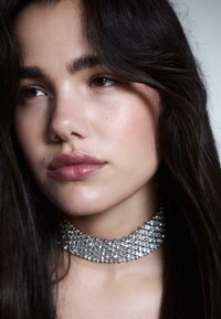 Choker-Halskette mit mehreren Reihen von funkelnden, klaren Kristallen, vor einem dunklen Hintergrund. Glatte, strahlende Haut und dunkles Haar umrahmen das Gesicht.