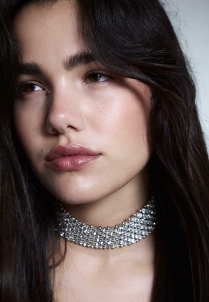 Choker-ketju, jossa on useita rivejä kimaltelevia kirkas kiteitä, tummalla taustalla. Sujuva, heloittava iho ja tumma hiuskehys kasvot.