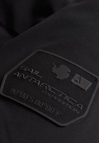 Svart tyg med en texturerad, rektangulär lapp som har "SAIL ANTARCTICA EXPEDITION," en karta över Antarktis och koordinater.