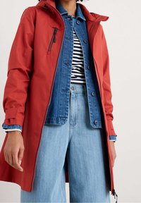 Rød vandafvisende jakke med hætte, der har en lynlåslomme foran, lagdelt over en blå denimskjorte og en stribet skjorte. Lyseblå jeans med vide ben.