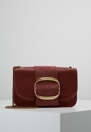 Sac bandoulière en cuir et daim rouge foncé avec grande boucle ovale dorée et anse chaîne dorée sur surface d'exposition blanche.
