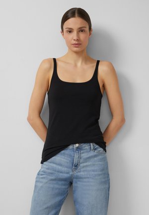 Femme portant un débardeur noir et un jean bleu clair, debout les mains derrière le dos contre un fond gris clair uni.