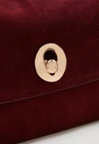 Burgundyröd mocka-clutch med ett cirkulärt klamp i guldton, med en slät textur och en ren design som framhäver klampen som en anmärkningsvärd detalj.