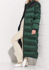 Długa zielona kurtka puchowa z czarnymi akcentami, zapinana na zamek błyskawiczny, z kieszeniami po bokach. Wykonana z błyszczącego, pikowanego materiału z wyraźnie zaznaczonymi panelami.