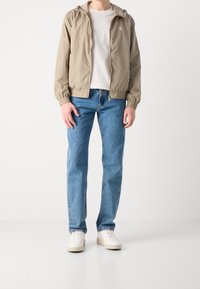 Lehká béžová bunda na zip s kapucí, nošená přes jednoduché bílé tričko; v kombinaci s modrými džíny a bílými teniskami.