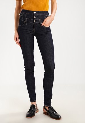 Femme portant un jean skinny foncé avec plusieurs boutons à l'avant, un haut côtelé moutarde et des chaussures noires à talon ouvert à lacets, debout, une main dans la poche.