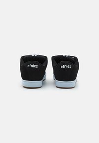 Baskets de skateboard noires au design bas profil, matériau semblable à du daim, logo "etnies" blanc sur le talon et semelle extérieure bleu clair.