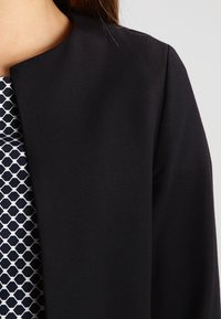 Svart jacka utan krage med slät textur, kombinerad med en marinblå och vit klänning med geometriskt mönster. Fokus på enkelhet och rena linjer.