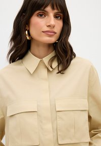 Camicia beige con colletto, caratterizzata da due tasche sul petto e una texture liscia. Chiusura con bottoni e polsini ripiegati dettagliando il design.