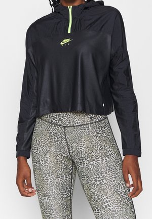 Osoba w czarnej krótkiej kurtce Nike Air z neonowym zamkiem i legginsach w panterkę, stojąca na jednolitym tle.