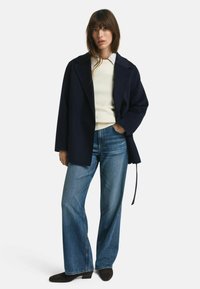 Marineblauwe jas met een relaxte pasvorm en brede revers, gedragen over een crèmekleurige trui. Gecombineerd met lichtblauwe wijde jeans en donkerbruine laarzen.