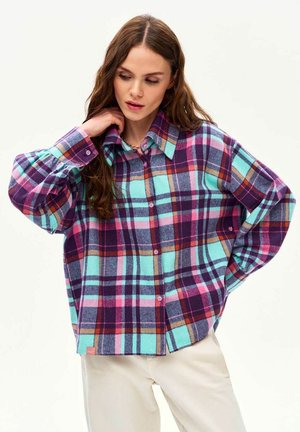 Kobieta w za dużej wielokolorowej koszuli w kratę w odcieniach fioletu, zieleni, różu i pomarańczy, ubrana w beżowe spodnie, pozująca z jedną ręką na kołnierzu, a drugą na biodrze.