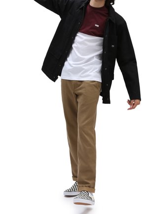 Personne portant une veste noire, un t-shirt Vans bordeaux et blanc, un pantalon kaki et des chaussures à enfiler à carreaux noirs et blancs en train de marcher.