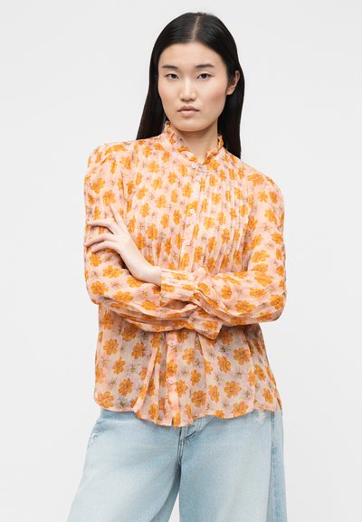 Γυναίκα με μακριά μαύρα μαλλιά φοράει ροζ μπλούζα με πορτοκαλί λουλουδάτο σχέδιο και ανοιχτό μπλε τζιν, στέκεται με τα χέρια σταυρωμένα μπροστά σε λευκό φόντο.