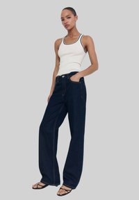 Weißes geripptes Tanktop mit schwarzem Rand, kombiniert mit dunkelblauen weit geschnittenen Jeans mit sichtbaren Nähten und einer Fronttasche. Schwarze Riemchensandalen.