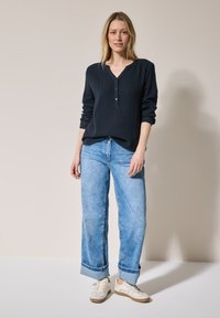 Camicia a maniche lunghe blu navy in tessuto con collo henley e dettagli in bottone, abbinata a jeans a gamba larga azzurri e sneakers bianche.