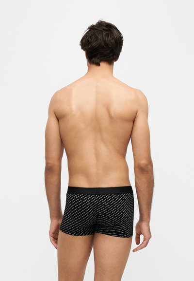 Homme debout, dos à la caméra, portant un boxer noir à motifs sur un fond clair uni.