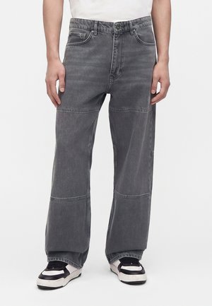 Person trägt locker sitzende, ausgebleichte schwarze Jeans mit sichtbaren Nahtdetails über den Knien und schwarz-weiße Turnschuhe.