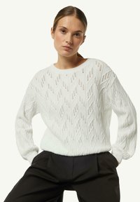 White knitted sweater with diamond-shaped patterns, crew neck, and ribbed cuffs, paired with black high-waisted pants.