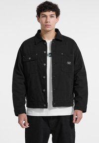 Chaqueta de mezclilla negra con cierre de botones, dos bolsillos en el pecho y un ajuste relajado. Combinada con una camisa blanca por debajo.