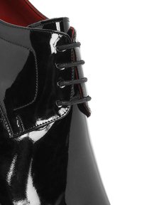 Chaussure en cuir verni noir avec une surface lisse et brillante, présentant un design à lacets plats et des détails intérieurs rouges.