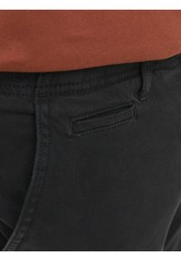 Černé bavlněné kalhoty s plochými kapsami, plochými švy a hladkou texturou. Část hnědého topu viditelná nahoře.