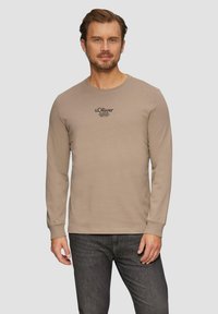 Beige lange mouw t-shirt van katoen, met een ronde halslijn en een zwart logo op de borst. Gecombineerd met donkere jeans.