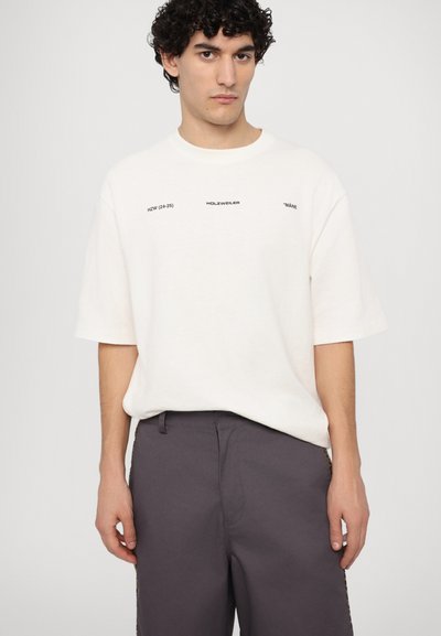 White cotton t-shirt with short sleeves, featuring black text and graphic prints. Paired with dark gray trousers, showcasing a relaxed fit.