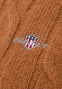 Pull marron tricoté avec un motif torsadé et un logo écusson brodé en rouge, blanc et bleu. Texture douce.