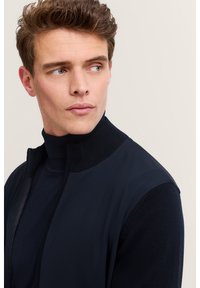 Granatowa kamizelka na zamek z wysokim kołnierzem, w połączeniu z ciemnym golfem. Posiada gładki materiał i dopasowany krój.