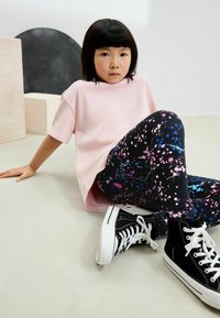Ljusrosa t-shirt med korta ärmar tillsammans med svarta leggings med ett färgglatt splattermönster och svarta högskurna sneakers.