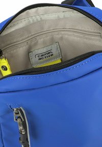 Bolsa de tela azul con cierre de cremallera; el interior presenta un forro beige, un parche con el logo y una correa de acento amarilla. Hardware de cremallera visible.