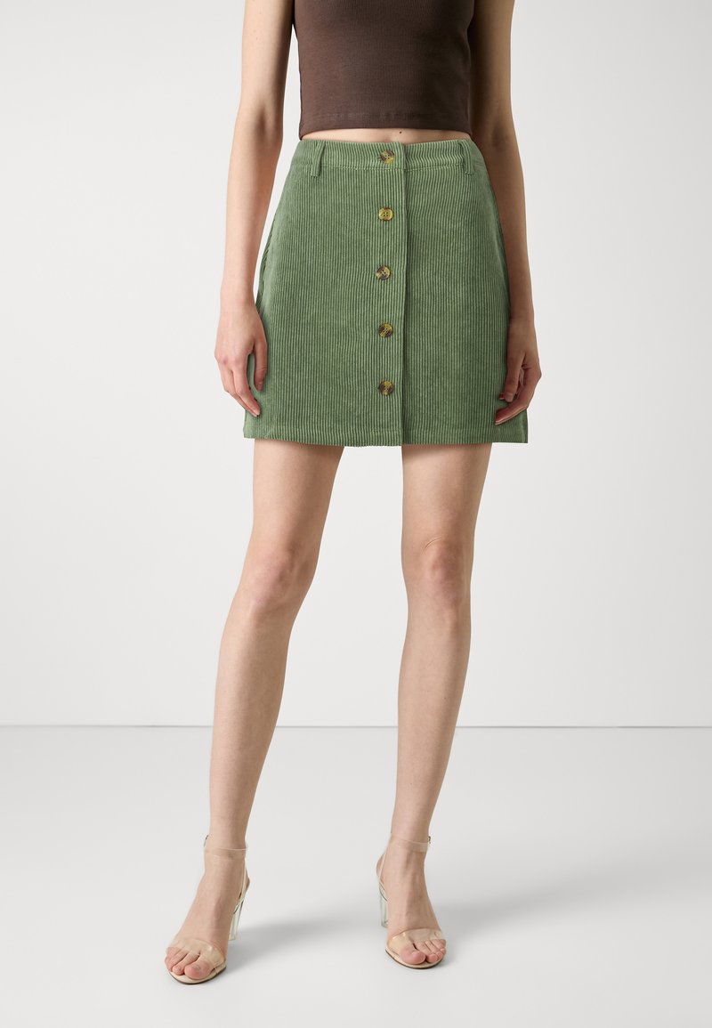 Mini-jupe en velours côtelé vert avec boutons sur le devant, tissu texturé et coupe droite. Portée avec un top marron ajusté et court, ainsi que des talons transparents.