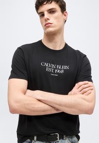 Νεαρός άνδρας με σταυρωμένα τα χέρια, ντυμένος με μαύρο t-shirt Calvin Klein που γράφει "CALVIN KLEIN EST. 1968" μπροστά, με απλό φόντο.